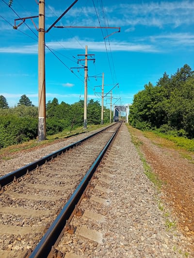 The image shows a set of railway tracks leading towards a bridge in the distance. The tracks are surrounded by ballast and are bordered by lush green trees and vegetation on either side. Several tall utility poles, likely for carrying electrical wires, are visible along the tracks. The sky is a clear blue with scattered white clouds. The scene appears to be in a rural or semi-rural area. The location is identified as Kolomak, Ukraine. No people or specific actions are depicted.