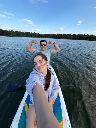 The image shows two people on a paddleboard in a body of water, likely a lake, given the presence of trees along the shoreline. A man, wearing sunglasses and a light-colored t-shirt, is flexing his bicep muscles for the camera. A woman, wearing a gray hoodie, is holding the camera and looking at the lens. A paddle lies on the paddleboard next to the woman. The sky is blue with some clouds. The location cannot be determined from the image.