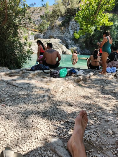 The image depicts a natural pool area with people engaged in various activities. Several individuals are visible in and around the turquoise water, with some wading and others preparing to enter. The surrounding environment features rocky terrain, lush greenery, and a small waterfall in the background. A foot is prominently in the foreground, suggesting a first-person perspective or a deliberate framing of the scene. The presence of a waterfall and the natural setting suggest the location is a popular spot for outdoor recreation. This scene is consistent with the natural bathing areas found near Ontinyent, Spain, such as the Pou Clar, known for its clear waters and rocky formations.