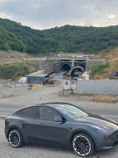 A dark gray Tesla Model Y is parked on a gravel surface in front of a large construction site. The construction site features unfinished tunnel entrances carved into a hillside covered in green trees and vegetation. There are concrete structures, scaffolding, and various construction equipment visible at the site. Orange traffic cones and yellow barriers are placed along the road and within the construction area. The sky is overcast with some patches of lighter clouds. The location is identified as Muğanlı, Azerbaijan. There are no people visible in the image.