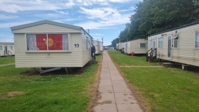 The image depicts a row of static caravans in a caravan park, likely located in Auchenblae, United Kingdom, given the context. A paved walkway runs between the caravans and the grassy area. In the foreground, the left side of the image shows the side of a caravan with the number '97' painted on it. A Kyrgyz flag is prominently displayed in the window of this caravan. Further down the row, another person is visible by the entrance of a caravan, partially obscured. Other caravans stretch into the distance, some with red gas cylinders outside. The environment appears to be a sunny day with a cloudy sky and lush green grass.