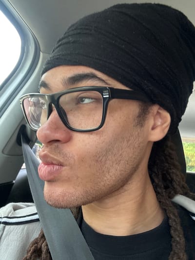 The media file captures a close-up portrait of a person inside a vehicle, likely a car, in Orsay, France. The image is oriented sideways, with the person's head tilted.

The subject is a person with light to medium skin tone, wearing a black head covering that appears to be a turban or durag. They have black-framed glasses resting on their nose, revealing a thoughtful gaze directed slightly upwards and to the left. Fine stubble or short facial hair covers their cheeks and chin. Brown, braided or dreadlocked hair is visible peeking out from under the head covering behind their right ear and extending towards the back of the neck. They are wearing a black shirt or undershirt, and a light-colored top (possibly a sweatshirt or hoodie) with a white and black striped detail on the sleeve is visible across their chest.

A grey seatbelt crosses their body, suggesting they are either a passenger or a driver in a stationary or moving vehicle. The car's interior, including part of the window frame and headliner, is visible in the background. Natural daylight streams in from the window, indicating it is daytime. The person's expression is neutral, showing no strong emotion. There is no visible text in the image.