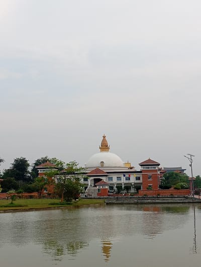 The scene captures a wide view of the Great Stupa and its surrounding structures within Lumbini Sanskritik, Nepal, under a uniformly overcast, light gray sky. A large, pristine white hemispherical stupa, topped with a golden, multi-tiered spire featuring a distinct finial, dominates the midground. This central edifice is integrated into a multi-story building complex with white lower sections displaying rectangular windows and several national flags (red, blue, yellow) on poles. The upper portions of the surrounding buildings feature red-brick construction, including prominent square towers with dark window bands and brown-tiled roofs. A smaller, ornate structure is partially visible to the far left, and another building with a traditional dark tiled roof is seen on the far right, partially obscured by dense green trees. In the immediate foreground, a calm body of water exhibits subtle ripples, reflecting the sky and the distant architecture. A strip of manicured green lawn, bordered by a low brick wall, separates the water from the main complex. Sparse, indistinct human figures are visible near the building's base and on the lawn. A tall, dark streetlamp equipped with a solar panel stands on the far right.
