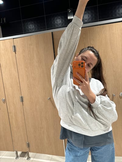 A woman takes a selfie in a public restroom. She wears a gray sweatshirt, blue pants, and a phone with an orange case. The background features wooden bathroom stalls with metal hardware and dark tiled walls. She holds the phone with her right hand, extending her left arm for the selfie. The photo is taken from an elevated angle.