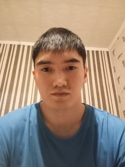 A young East Asian male, appearing to be in his late teens or early twenties, is pictured from the shoulders up, looking directly at the camera. He has short, dark hair and dark eyes. He is wearing a plain, light blue t-shirt. The background consists of striped wallpaper in shades of gray and white, with a white ceiling above. The lighting appears to be artificial, coming from above and slightly in front of the subject. The image was taken indoors, in what appears to be a residential setting. No specific location in the city of Khutorok, Russia can be determined from the visual information provided.