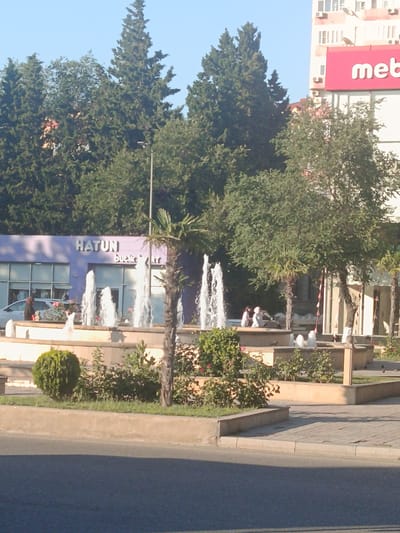 The image depicts an outdoor scene with a fountain in the foreground. Several people are visible near the fountain and in front of a building with the name "HATUNI" and "Buckle & rt" visible. The building appears to be a store or business. In the background, there are trees and a tall building with a prominent red sign that reads "meb". The sky is clear and blue, suggesting it is daytime. The presence of fountains and manicured greenery indicates a public space or plaza. The overall atmosphere appears to be relaxed and casual.