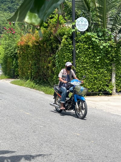 A person wearing a face mask and a cap rides a blue motorcycle down a paved road. The person is wearing a tan t-shirt, blue jeans, and a plaid scarf draped over their shoulders. The motorcycle has a basket on the front. Behind the motorcycle, there is a tall, dense green hedge. To the left of the hedge, there is a sign with a house logo and text that reads "Baan Tusneem" and a phone number, "093 646 7871". Further to the left, there are trees with red flowers. The road is surrounded by greenery, suggesting a rural or suburban setting. The sun is shining, casting shadows on the road. The image was taken in Choeng Thale, Thailand.