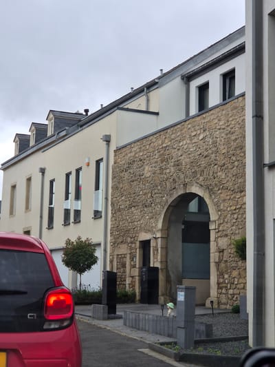 The image shows a street scene in Filsdorf, Luxembourg, with a red car in the foreground. The car, a Citroën C1, has its brake lights illuminated. Behind the car is a building with a beige facade and several windows. The building features a stone-clad section with an arched entryway. To the left of the stone section, there is a small tree and some shrubbery. Further down the street, a modern building with a grey exterior is visible. The sky is overcast.