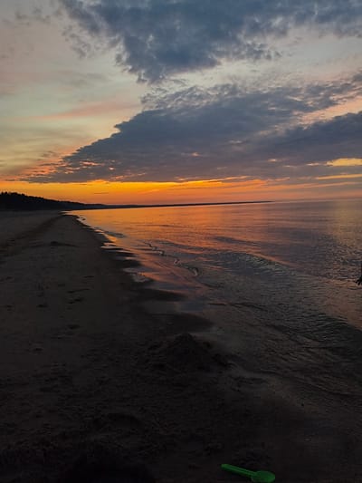 A wide shot captures a beach scene at sunset in Apšuciems, Latvia. The sky is filled with dramatic clouds, transitioning from a bright orange horizon to darker grays and blues overhead. The ocean stretches out to the right, reflecting the warm hues of the sunset. The foreground consists of a sandy beach with footprints visible, leading towards a dark tree line in the distance. A small green plastic shovel rests on the sand near the water's edge. There are no people clearly visible in the frame.