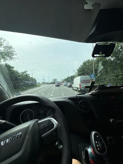 Here is a forensic description of the image:

The image is taken from the interior of a vehicle, likely a truck due to the "IVECO" logo visible on the steering wheel. The vehicle is in traffic, stopped behind a white van with red chevron markings on the back. The gear shift is in the park position (P). The speedometer indicates the vehicle is stationary. The image shows the view through the windshield.  The location is Houthalen, Belgium, indicated by the road sign in front of the van.  The environment appears to be an urban road with trees lining the sides. It is daytime, and the sky is overcast. The windshield shows water droplets.