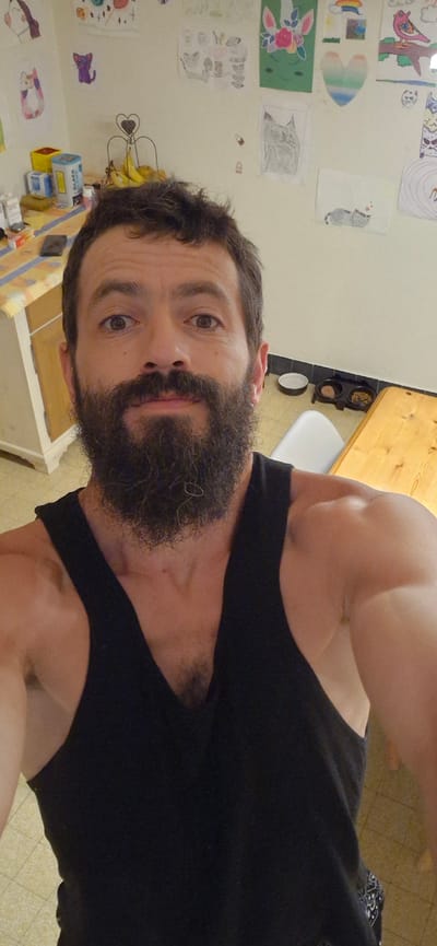 A man with a beard and dark hair, wearing a black tank top, poses for a selfie in what appears to be a domestic setting. The background features a wall adorned with various children's drawings, including cats, a unicorn, and a rainbow. To the left, a kitchen counter is visible with several boxes and a bunch of bananas in a stand. To the right, a wooden table and two pet bowls can be seen. The floor is tiled. The man's left arm and shoulder are prominent in the foreground, with his right shoulder also partially visible. The overall lighting suggests an indoor environment. The image is a close-up, self-taken photograph. There is no information in the image to suggest this location is in Wattenwil, Switzerland.