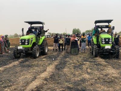 The image shows a group of people gathered in a field in Ringimawa, Nigeria, at coordinates 11.666 latitude and 8.416 longitude. The field is covered in recently burned grass. There are two green tractors, both labeled "WOFAN," parked in the field. The tractors are facing each other with a group of people standing between them. Several people are in front of each tractor, with a few more people standing at the back of the field. The people are a mix of men and women, some wearing traditional clothing. A few people are holding their hands up, possibly waving. It appears the group of people are gathered to see the tractors.