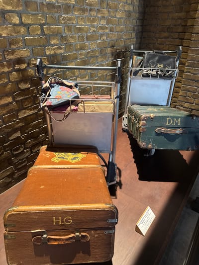 This image depicts a staged scene resembling Platform 9 3/4 from the Harry Potter series. Two luggage trolleys are loaded with vintage suitcases bearing the initials "D.M." and "H.G.," alluding to characters Draco Malfoy and Hermione Granger. One trolley also has a black bag and the other has a floral backpack. A small box labeled "HERMIONE GRANGER'S TROLLEY CART" in Japanese is present, suggesting this is part of an exhibit. The scene is set on a stone-brick floor, with a wood-paneled wall and floor in the background. The location is implied by the text on the box.
