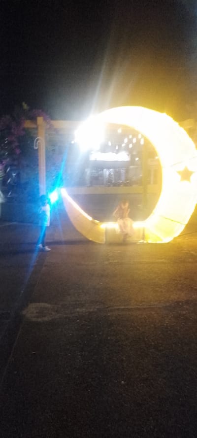 The image displays a nighttime scene in the city of La Fría, Venezuela. In the foreground, a dark, paved surface occupies the lower half of the frame. The central subject is a large, illuminated crescent moon structure, glowing brightly with a warm yellow-white light, and featuring a distinct star shape on its upper right curve. A person is seated on the lower interior edge of this moon structure. To the left, another individual stands, largely obscured by an intense blue light source originating near them. In the mid-ground, behind the illuminated moon, structural elements of what appears to be a building or market stall are visible, adorned with multiple small lights. To the upper left, a wooden archway with an overhead crossbeam is present, with purple flowering foliage behind it. The overall environment is dark, illuminated by the prominent artificial lights of the moon structure and the blue light source.