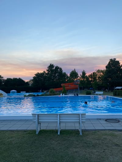The image depicts an outdoor swimming pool area in Deutschkreutz, Austria, during dusk. The sky displays a gradient of pale blue to soft pink and orange hues from the setting sun. In the foreground, a light-colored metal bench with horizontal slats sits on a patch of green grass. Behind the bench, a large, bright blue swimming pool occupies the majority of the frame. The water's surface is slightly rippled, reflecting the colors of the sky.
Further back, on the left side of the pool, a white structure with a yellow accent is visible, possibly part of a water feature or slide. In the center-left, an orange water slide with two distinct chutes is prominent, set against a backdrop of dark green trees. Several people are present in and around the pool area, with some swimming and others socializing or walking in the background. The perimeter of the pool is lined with light-colored square tiles. Trees form a dense, dark green border around the swimming pool complex. The overall atmosphere suggests a relaxed evening at a recreational facility.
