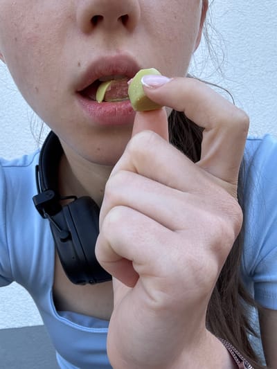 This is a close-up, vertically oriented, full-body shot of a person in Erlenbach am Main, Germany. The person is wearing a light blue t-shirt and black headphones. Their right hand is visible, with painted fingernails, holding a small, light green, soft candy or gummy to their open mouth. Their lips are red and slightly parted, revealing their tongue. The background is a plain white surface, possibly a wall or backdrop. The lighting is bright, suggesting daytime, but there are no clear indicators of the weather or a specific location within Erlenbach am Main. The focus is on the person's hand and mouth, with the candy poised to be eaten. There is no visible text in the image.
