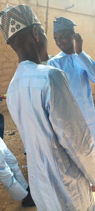 The scene, located in Ungogo, Nigeria, features three male individuals outdoors. The primary subject in the foreground is observed from the rear-right profile, wearing a light blue, long-sleeved traditional garment with subtle vertical stripes and dot patterns, and a beige cap adorned with blue, orange, and black geometric embroidery. To his right and slightly behind, a second male subject is visible from the chest up, facing forward with his head slightly turned left, displaying a smile. He wears a similar light blue garment and a dark blue cap with light blue horizontal stripes, which he is adjusting with his right hand near his ear. In the bottom-left corner, the lower legs and feet of a third individual are visible, clad in light blue trousers and black shoes, positioned on dry, reddish-brown earth. The background consists of a light brown, textured wall, likely constructed from mud or unpainted brick. The environment is under direct natural sunlight, creating distinct shadows.