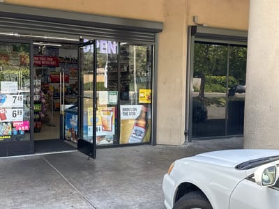 The image shows the brightly lit entrance of a convenience store or liquor store in North Highlands, United States, on a clear day. The storefront features large glass doors and windows, with a security shutter installed above. One of the glass doors is open, revealing the interior of the store.
Inside, shelves stocked with various merchandise, including snacks and beverages, are visible. Display refrigerators, one notably branded "Foster Farms Dairies," are also present. A neon "OPEN" sign is partially visible in the reflection.
Promotional signs and posters adorn the windows. A large red banner inside reads, "THANK YOU FOR SHOPPING WITH US!" Handwritten signs on the left door advertise "Milk 1/2 gal 2 for $6" and "1 gal 2 for $10," with prices "$7.49" and "$6.89" displayed nearby. A pink sign indicates "poppi Sold Here." On the right window, a large Budweiser poster features a bottle and the text "BRING ON THE BUD." Another Budweiser poster states "THIS BUD'S FOR YOU." Other signs include "24/7," "AM CA," "RED 100'S BOX," and "$8.64," likely for tobacco products. A green "PASS" sign and a yellow "EARN GROW REDEEM SPIRIT" sign suggest loyalty programs.
The concrete ground in front of the store shows some wear. Reflections of trees and a black car are visible in the glass panels to the right of the entrance. The hood and front wheel of a white car are visible in the foreground, parked close to the store entrance. The building exterior features a light-colored, textured stucco finish, with a large, round pillar on the far right. No people are visible, and no specific activities beyond the store being open are depicted.
