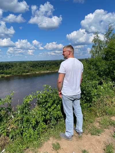 A man with short blonde hair and glasses stands on a grassy hill overlooking a wide river. He is wearing a white t-shirt and light blue jeans, with his back to the camera. The river is dark and reflects the blue sky, which is dotted with fluffy white clouds. Lush green trees and vegetation line the riverbank and the hill where the man is standing. The background shows a dense forest stretching to the horizon. The scene appears to be a natural landscape with no visible buildings or urban elements, suggesting a location outside of the city center of Kirovo-Chepetsk, Russia, possibly in a scenic natural park or rural area.