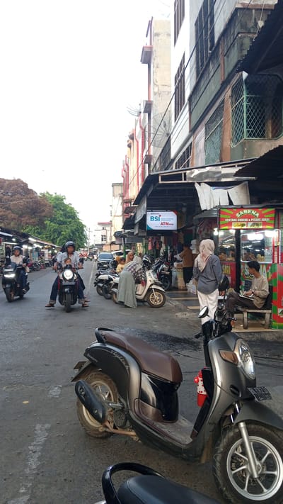 The image depicts a street scene in Puloara, Indonesia. A road is lined with shops, some with awnings extending over the sidewalk. Several motorcycles are parked and in motion on the street. In the foreground, two scooters are visible, one is black and grey with a brown seat, and the other is partially visible behind it. On the street, a man wearing a helmet is riding a motorcycle with a child seated in front of him. To the right, a woman wearing a headscarf and a long skirt is leaning over a white scooter. Another woman in a grey headscarf and white pants is walking away from the camera. Behind them, a man is sitting on a bench, looking at a book or paper. A sign for "BSI BANK SYARIAH ATM MINI" is visible on a building facade. Further down the street, more people and motorcycles are present, and market stalls are set up. Trees are visible in the background on the left side of the image. The sky is overcast.