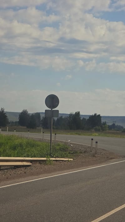 The image depicts a rural roadside scene under a partly cloudy sky. A paved road with a white dividing line curves to the right. Along the edge of the road, there is a gravel shoulder and a metal guardrail. A traffic signpost with two signs, a circular one above a rectangular one, stands to the left of the road. Beyond the road, there is a grassy embankment and a line of trees forming a horizon. In the distance, rolling hills and a hazy landscape are visible. The overall environment suggests a quiet, open area outside of a densely populated urban center.