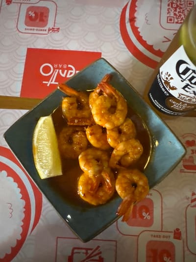 This is a top-down, slightly angled shot of a dish of cooked shrimp and a slice of lemon on a blue square plate. The shrimp are coated in a glossy, reddish-brown sauce and are arranged in a circular pattern. A slice of lemon sits at the bottom of the plate. In the background, a bottle of Corona beer is visible, along with some red and white patterned paper coasters or placemats. One of the coasters has text that appears to be in Spanish, including "CERVEZA MODELO." Other visible text includes "BENTO-DRIVE," "TAKE OUT," and percentage symbols, suggesting a takeout or casual dining context. The lighting is bright, indicating it could be daytime. There are no people visible in the image. The overall scene suggests a meal that has been served, possibly at a restaurant or for takeout. The location context of Kryzhanivka, Ukraine, doesn't seem to be directly evident from the visual cues within the image itself.