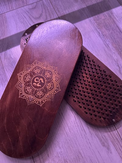 Two wooden sadhu boards are placed on a wooden floor with a purple light cast over the scene. One board, in the foreground and on the left, is smooth and features a laser-etched mandala design with the symbol "Om" in the center. The second board, to the right and slightly behind, is also made of wood and has rows of metal nails protruding from its surface. The floorboards are oriented diagonally in the image. No people are present in the image. The location is described as Высокая Гора, Russia.