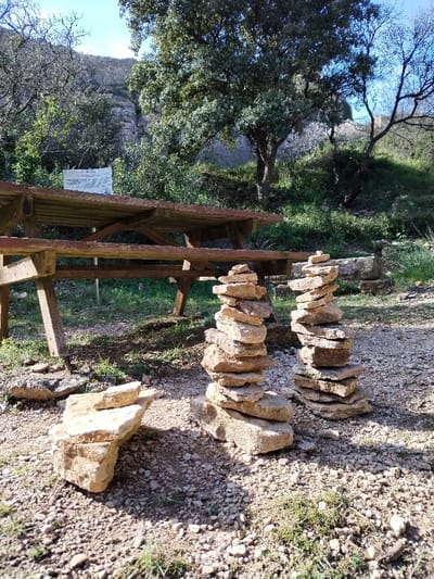 This image captures an outdoor scene, likely a picnic or rest area in a natural setting, under bright daylight. The ground in the foreground and midground is composed of light-colored gravel and small stones, interspersed with patches of green grass.
In the immediate foreground, to the left, is an informal pile of several light brown, irregularly shaped stones. Slightly further back and to the right, are two prominent cairns, or stacks of flat, light brown stones, carefully balanced one on top of the other, forming tall, slender columns. The cairn on the left appears slightly taller and wider than the one on the right.
Dominating the midground is a sturdy wooden picnic table with attached benches, made of thick, weathered planks. Its construction features diagonal supports beneath the tabletop and benches. The table is oriented diagonally to the left, with its length extending towards the left side of the frame.
Behind the picnic table, to the left, a white sign is visible. It appears to have black text or symbols on it, but the details are too small and blurred to be legible. The sign is mounted on a post or frame, slightly obscured by surrounding foliage.
The background consists of dense vegetation and hilly terrain. A variety of trees with green leaves and some with bare branches are visible, suggesting a mix of deciduous and evergreen species. The foliage is lush and green, indicating a healthy natural environment. Behind the trees and vegetation, a rocky or mountainous slope rises, covered in sparse greenery. The sky above is a clear, bright blue, suggesting good weather conditions. Sunlight is strong, creating distinct shadows on the ground and highlighting the textures of the stones and wood. The overall impression is one of a serene and natural outdoor space, possibly a trailside stop or a designated recreation area.