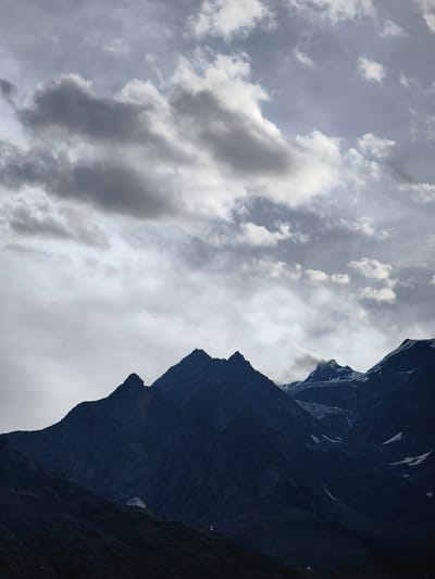 The image shows a dramatic mountain landscape under a cloudy sky. The foreground features a dark, silhouetted mountain range with a small white building nestled on a grassy slope on the left side. The building appears to be a simple, rectangular structure with a dark roof. Further back, the peaks of other mountains are visible, some with snow-capped summits and glaciers. The sky is filled with a mix of dark, heavy clouds and lighter, more wispy formations. The overall atmosphere is moody and serene, with the light suggesting either early morning or late afternoon. The location is identified as Gushal, India.