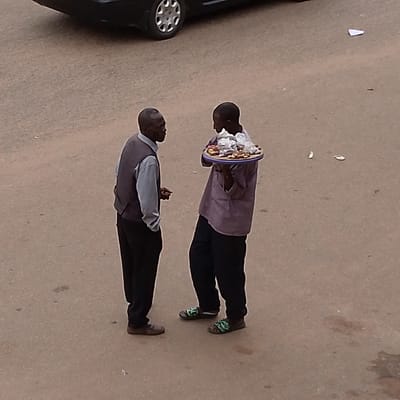 Two men are standing on a paved surface, possibly a street or sidewalk. The man on the left is wearing a grey vest over a light blue collared shirt and dark pants. He has his hands clasped in front of him. The man on the right is wearing a purple shirt and dark pants. He is holding a circular tray in front of him, which appears to contain various food items, some wrapped in plastic. The background shows a black car with its wheel visible, and the ground has some debris scattered on it. The lighting suggests it is daytime. This scene is not in Kayseri, Turkey.