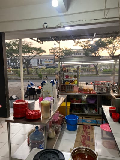 The image shows an outdoor food stall in Mekar Bakti, Indonesia, at dusk. The stall is set up on a tiled floor and is enclosed by a metal structure with a roof. In the foreground, there are stacks of red and white plates, as well as various plastic containers and bottles of drinks. A large blue water jug is visible on a lower shelf.

Behind the stall, a street is visible with trees and buildings lining the road. A blue motorcycle is parked on the side of the street, near a table and chairs. The sky is orange and yellow, indicating the setting sun. A security camera is mounted on the ceiling above the stall. The overall impression is of a typical street-side food vendor preparing for or concluding their business day.