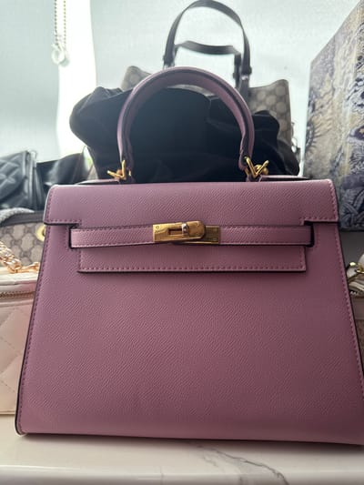 A pink leather handbag with gold hardware is displayed prominently in the foreground. The handbag has a structured shape with a top handle and a flap closure secured by a gold turn-lock mechanism. The texture of the leather is visible, showing a fine pebble grain. Behind the pink handbag, other bags are partially visible. To the left, a black quilted handbag and a grey and beige patterned handbag are present. To the right, a portion of a patterned fabric item, possibly another bag or textile, is seen. The scene appears to be an indoor setting, possibly a retail environment or a personal collection display. There are no people visible in the image. The lighting suggests an overhead source, casting subtle shadows and highlighting the textures of the bags. The background is out of focus, emphasizing the main subject.