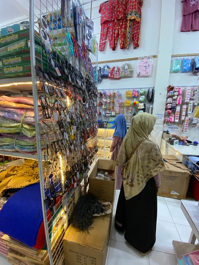 This is an eye-level, indoor shot of a store aisle filled with merchandise. A person, likely a woman based on the hijab and clothing, is seen from the back, wearing a light brown hijab and a black garment. She appears to be browsing or shopping. The aisle is lined with shelves displaying a variety of items. The shelves on the left hold colorful fabrics or clothing, possibly garments like sarongs or dresses, folded neatly. The shelves on the right are packed with rows of eyeglasses, with many pairs displayed on racks. Some of the eyeglasses have price tags attached. In the background, more shelves hold various small items, possibly accessories or toys, including what look like keychains with animal figures. Cardboard boxes are visible on the floor and on some of the shelves. One box has text that includes "Measurement 760 x 600 x 780 mm" and "GW: 20 PCS N.W.: 13 KGS MEAS: 56.5x35.5x36.5CM". The lighting suggests it is daytime or that the store is well-lit. The overall impression is of a busy retail environment, likely a small shop or market stall, possibly in Indonesia given the location context. The scene conveys a sense of everyday commerce.