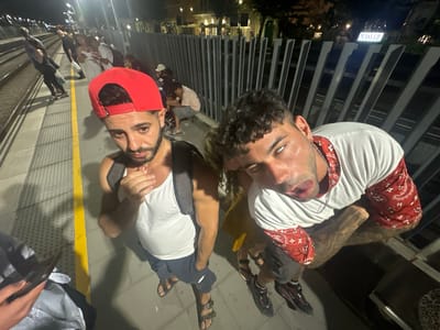 At night, on a train platform in El Masnou, Spain, two young men are visible in the foreground. The man on the left, wearing a red baseball cap, white tank top, and dark shorts, is looking down and slightly to the left. The man on the right, with curly hair, is looking directly at the camera with his mouth slightly open, wearing a white t-shirt with red bandana-patterned sleeves and dark shorts. They are standing next to each other, with the man on the right appearing to be leaning slightly.
In the background, several other individuals are visible, some standing and others sitting. One person in the middle ground is wearing a white dress. The platform is bordered by a yellow line and a metal fence. Beyond the fence, there are buildings with lights and a sign that reads "MASTER". The scene suggests a social gathering or a moment of leisure in a public space.