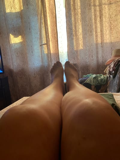 The image is a first-person perspective shot of a person's legs and feet lying on a bed. The legs are bare and appear to be tanned. The feet are visible at the top of the frame, with toes pointing upwards. The background consists of a patterned curtain illuminated by sunlight, creating a warm glow and casting shadows. To the right, a pile of clothes and a straw hat are visible on a dresser. The scene is indoors, in a bedroom. There is no information in the image or the provided context that allows for the determination of the location in the city of Podkletnoe, Russia.