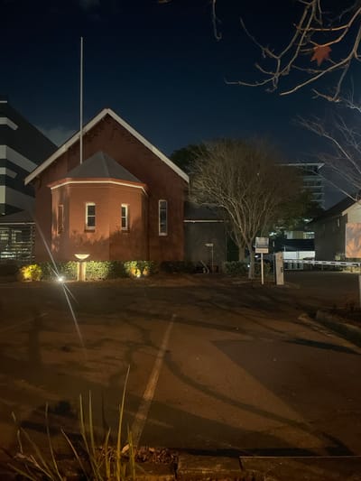 The image depicts the exterior of a small, red brick building with a distinctive octagonal projection at the front, illuminated by ambient lighting at night. A tall flagpole stands against the dark sky, positioned near the peak of the building's roof. The building appears to be part of a larger complex, with a modern, multi-story structure visible to the left, characterized by its dark facade and white horizontal accents. To the right, a gate with a barrier arm is visible, suggesting a controlled access point to the property. Bare trees with spindly branches are scattered throughout the scene, indicating a cooler season. The ground in front of the building is a paved area, possibly a parking lot, with visible white road markings and shadows cast by the lights and structures. In the foreground, some blades of grass and fallen leaves are visible along the edge of the paved area. The overall atmosphere is one of quietness and stillness, typical of a nighttime urban environment. The building's architectural style and the surrounding modern construction suggest it could be a historical building preserved within a contemporary urban setting in Fortitude Valley, Australia.