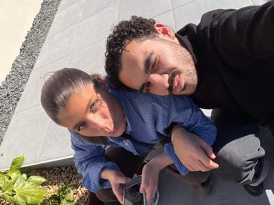 This image captures two individuals, an adult female and an adult male, in an outdoor setting in Marche-lez-Écaussinnes, Belgium. Both subjects are crouched or kneeling, positioned closely together and looking upwards towards the camera. The scene is brightly lit by direct sunlight.

The female, positioned on the left side of the frame, has dark hair pulled back and blue eyes; she wears a blue and white checkered long-sleeved shirt and dark trousers. She holds a smartphone displaying an image on its screen in her left hand. Her right shoulder is partially covered by the male subject's head.

The male, situated on the right, has dark, curly hair and a beard. He wears a black long-sleeved hooded top, dark grey pants, and black shoes. His head rests on the female's right shoulder, and his left hand, adorned with a thin bracelet, rests on her right arm. Both individuals have their lips slightly pursed, and the male's right eye is partially closed.

The environment includes a light gray tiled surface covering most of the ground directly beneath the subjects. To the left, there is a border of dark grey gravel, and further left, an area of wood chip mulch with green foliage, including a plant with large, light green leaves, is visible in the lower-left corner.