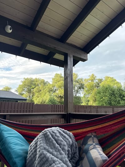 The image depicts a hammock with colorful stripes, adorned with a teal pillow and a gray blanket. A patterned pillow is also visible. The hammock is situated on a wooden deck under a wooden roof, which has string lights attached to the underside. A small white security camera is mounted on one of the wooden beams. In the background, a brown wooden fence encloses a yard with lush green trees and a cloudy sky overhead. To the left, a dark-colored roof of a building is partially visible. The scene appears to be located in the city of Gavshino, Russia, suggesting an outdoor recreational space.