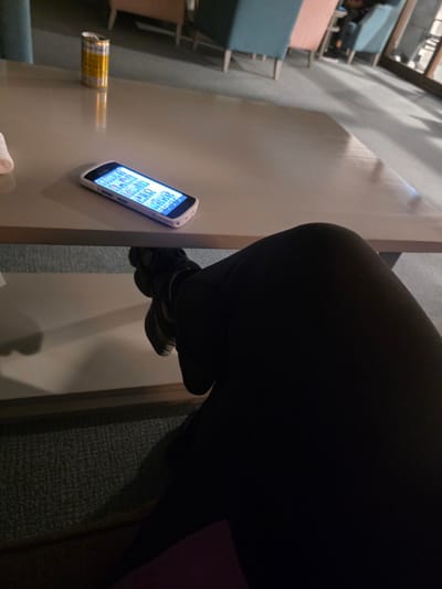 The image shows a table with a mobile phone and a can. A person wearing black pants is sitting at the table. The location is potentially Harrogate, United Kingdom, but this cannot be verified from the image.