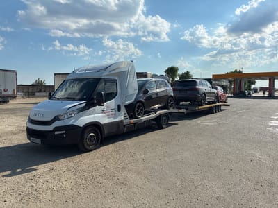 An Iveco car carrier truck is parked on a gravel lot under a partly cloudy blue sky. The truck is white with the Iveco logo visible on the front. The truck bed is loaded with two cars: a dark grey hatchback and a dark grey SUV. The hatchback is positioned on the lower deck of the carrier, and the SUV is on the upper deck. In the background, there is a gas station with an orange awning and several other vehicles. The ground is covered in loose gravel and some asphalt. The image appears to have been taken during the daytime.
