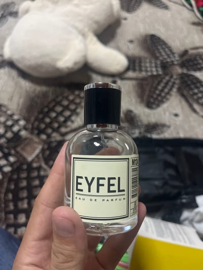 A close-up shot shows a person's hand holding a clear glass perfume bottle with a black cap. The bottle has a label with black text on a cream background. The text on the label reads "EYFEL EAU DE PARFUM" and some barcodes. The background is blurred but appears to be an indoor setting with patterned fabric. The overall lighting suggests it is daytime. There are no specific location cues beyond the general indoor setting. The image focuses on the product itself.