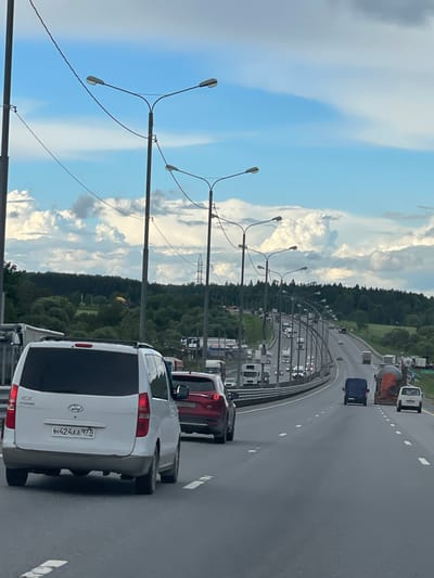 The image depicts a congested multi-lane highway in Бавыкино, Russia. Cars and trucks are densely packed, indicating heavy traffic. The sky is partly cloudy with a clear blue expanse. The roadside features a forested area punctuated by streetlights. An electrical transmission tower is visible beyond the trees. The rear of a white Hyundai Starex van is in the foreground, its license plate visible as Н424АА 977.