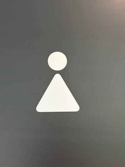 A white circular shape is positioned above a white triangular shape, forming a stylized human figure. This figure is centered on a dark grey, textured background. The image is a close-up, likely of a sign or decal. The specific location in Son en Breugel, Netherlands, is not discernible from the visual information provided.