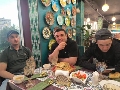 Three men are seated at a table in a restaurant, engaged in a meal. The man on the left wears a dark green polo shirt and a black baseball cap. He looks towards the camera with a neutral expression. The man in the center, wearing a black t-shirt, is looking at the camera and resting his chin on his hand, appearing relaxed. The man on the right, wearing a black jacket and a dark baseball cap, is looking down at a mobile phone in his hand.

The table is set with several dishes. In the foreground, there are two ornate glasses, one containing a dark liquid and the other empty. A small white bowl with a light-colored liquid, likely tea, is also present. A basket of bread is in the center of the table, next to a white napkin holder. A dish of salad, containing tomatoes and cucumbers, is to the right of the bread. There are also plates and gold cutlery on the table.

The restaurant's wall is decorated with numerous colorful plates, featuring geometric patterns and depictions of fruits and buildings. Green patterned fabric hangs on the wall behind the man on the left. The ceiling is dark, with several modern pendant lights providing illumination. Lush green plants are visible in the background, suggesting an indoor dining environment. Through a window on the left, an urban landscape with buildings and trees can be seen, indicating it is daytime. The overall atmosphere appears casual and relaxed, suggesting a social gathering. No visible text can be deciphered from the image.