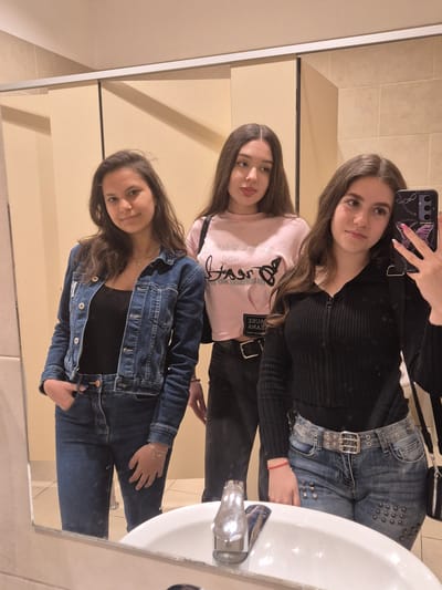 Three young women are captured in a mirror selfie, likely taken in a bathroom in Zagore, Bulgaria. The image is oriented vertically but the subjects appear to be standing upright.
The woman on the left, holding the smartphone, has long brown hair and is wearing a black ribbed long-sleeved top. She is also wearing dark jeans with metal studs or grommets and a wide, transparent belt with multiple silver buckles. Her phone case features a purple butterfly and chain design.
The middle woman has long brown hair and is dressed in a light pink long-sleeved top. Visible text on her shirt, when seen reflected, reads "NO CAUSE DREAMS" in a stylized script. She is wearing dark pants or jeans, and a black strap from a bag or belt displays the mirrored text "CAUSE JEANS".
The woman on the right has long brown hair and is wearing a black top underneath a blue denim jacket with silver buttons. She is dressed in blue jeans and accessorizes with a silver necklace and a delicate silver chain bracelet on her left wrist.
All three women look towards the camera with casual expressions, suggesting a friendly interaction. The background shows light beige tiled walls, typical of a bathroom setting. A white ceramic sink and a chrome faucet are visible on the right side of the reflection. The lighting is artificial, indicating an indoor environment.