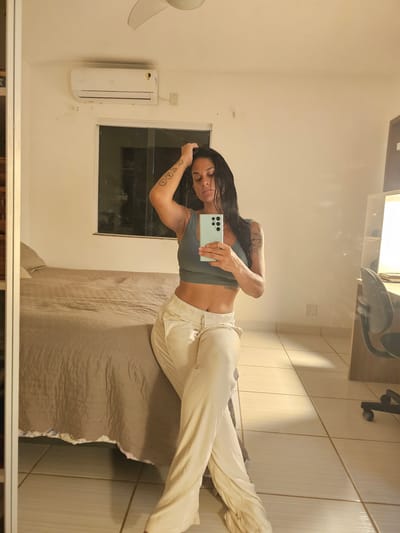 The image displays a mirror selfie of a woman in an indoor setting, likely a bedroom in Cabo Frio, Brazil. The photo is oriented sideways, but depicts the subject standing vertically.

The central figure is a woman with long, dark, slightly wet-looking hair. She is wearing a light grey or dusty blue ribbed crop top that reveals her midriff, and light-colored (possibly beige or off-white) relaxed-fit drawstring pants. She holds a light green smartphone, likely a Samsung Galaxy S series due to the distinctive camera module, in her right hand, focusing her gaze on the screen. Tattoos, including some circular symbols, are visible on her right forearm. Her overall posture and expression appear relaxed.

The reflection in the mirror reveals elements of the room. To the left of the woman, a white split-unit air conditioner is mounted on a light-colored wall, adjacent to a dark, rectangular window. Below her, a bed with a textured greyish-brown bedspread is partially visible. To her right, a section of wall covered with vertical light-colored tiles can be seen, with a reflection of a light fixture and what appears to be an older landline phone on a shelf above. The room is illuminated by artificial light, suggesting the photo was taken during the evening or night. No discernible text is visible on the woman's clothing or in the room, apart from the phone's design.