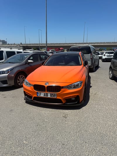 The image shows an outdoor parking lot filled with various vehicles under a clear blue sky, suggesting a sunny day. The perspective is from ground level, looking towards a row of parked cars.

The most prominent subject is a bright orange sports car, positioned in the center of the frame. It has a black grille and aggressive front-end styling. The car's license plate is visible and reads "17 AID 058 TR". To the left of the orange car, a silver hatchback is partially visible, and behind it, several other cars and vans are parked, including a white van and a dark-colored vehicle. In the background, a bridge or overpass can be seen with streetlights lining its edge. The ground is paved with asphalt.

The scene appears to be a typical parking area, possibly near a commercial or public space. The clear sky and bright sunlight indicate it is daytime, likely mid-morning or afternoon. There are no people visible in the image. The overall impression is one of a static scene of parked vehicles on a bright, clear day. The location context provided is Çanakkale Merkez, Turkey, which aligns with the Turkish license plate on the orange car.