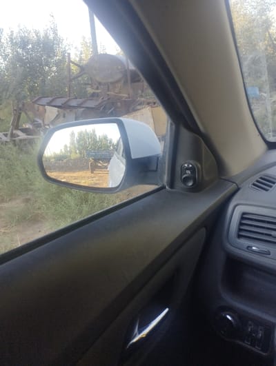 The image is a first-person view from inside a vehicle, looking out the driver's side window and into the side mirror. The vehicle appears to be parked in an outdoor, rural setting, with dirt ground and sparse vegetation. In the distance, there is a large, rusted piece of agricultural machinery.

The side mirror reflects a tractor with a blue trailer, parked on a dirt road. Trees are visible behind the tractor, and the sky is bright, suggesting it is daytime. The interior of the car is visible on the right side of the frame, showing part of the dashboard with an air vent and a window control button. The overall impression is of a somewhat derelict or unused area. The provided image does not contain any information that would allow for the specific identification of the location as Margabat, Uzbekistan.