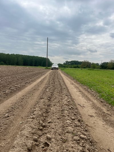 The image shows a dirt road or track running between a freshly plowed field on the left and a grassy field on the right. The road has distinct tire tracks, indicating it is used by vehicles. A white van is seen in the distance on the road. On the left side of the road, there is a line of trees and a utility pole with power lines running alongside the field. The sky is overcast with gray clouds. The location is speculated to be in the city of Tseretelevo, Bulgaria.