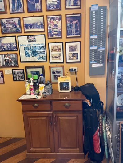 This detailed description pertains to an indoor scene in Ban Yid, Thailand. The focal point is a muted orange-ochre wall adorned with a dense grid of framed items, predominantly photographs, centered above a dark wooden cabinet. Most frames are dark brown, with one prominent black frame containing a newspaper clipping. The photographs depict various groups of people, frequently featuring motorcycles, suggesting a theme related to motorcycle enthusiasts. Styles range from vintage black-and-white to contemporary color images. The large black-framed item is a newspaper article from "Phuket Insider" with a headline that includes "Harley a single outlaw in sight." Accompanying images within the clipping show motorcycles and individuals. To the right of the framed items, a gray wall-mounted rack is visible, featuring fifty numbered slots (01-50). Several slots contain white or blue cards, some of which are larger pieces of paper covering multiple slots, indicating its use for time cards or identification. Below the wall display, a dark wooden cabinet stands on a tiled floor. The cabinet has two upper drawers, each with a brass-colored shell-shaped pull, and two lower doors with carved wooden handles. The top surface of the cabinet serves as a display area for various objects: * A transparent plastic cylinder containing white Yonex badminton shuttlecocks. * A pink bottle of "Kamin" brand lotion. * A green spray can (3M Leather & Vinyl Restorer). * A black spray can (CRC Rust Remover Foam). * A tall, slender brown bottle, possibly containing a lubricant. * A blue plastic bottle. * A white and orange pouch of "Shell Advance" motorcycle oil. * A gray bottle of "CASTROL V-TWIN CLUTCH OIL." * A small, chrome-finished cup or container. * A stack of papers with handwritten notes, secured by a blue pen. To the right of these items, a yellow "TimeTech KL-6500" analog clock displays the time as approximately 1:50. It is connected by a black power cord to a white electrical outlet on the wall. Leaning against the right side of the cabinet are a black, red, and white golf bag, from which an orange-tipped golf club is partially visible, and a cluster of four umbrellas. The umbrellas vary in design, including silver, green, and patterned fabric. The floor beneath the cabinet is tiled with a diagonal pattern of alternating light and dark brown wood-grain strips. To the far right, a vertical section of a glass display cabinet is visible, containing